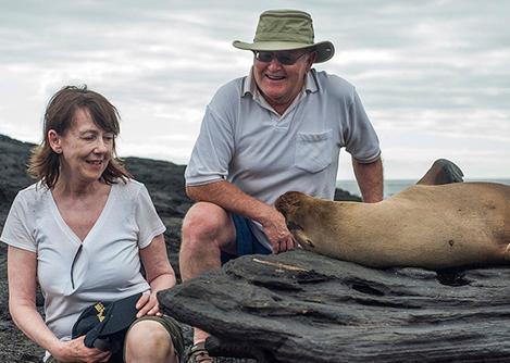 Smiling visitors sitting next to a playful sea lion on a rock formation in the Galapagos Islands.