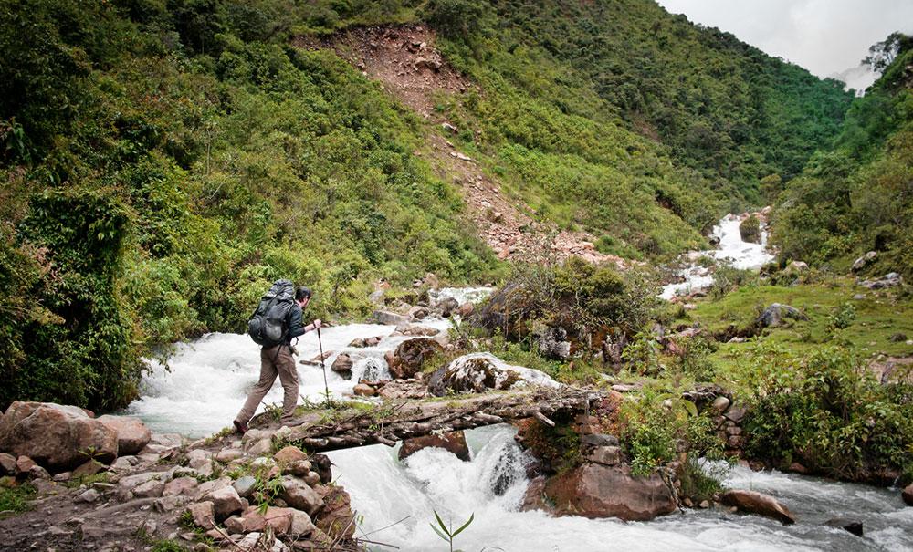 A man carrying hiking poles is walking over a bridge with water flowing under it. In front of him are mountains with green plants